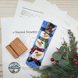May include: A fused glass art piece featuring three stacked white snowmen on a light blue rectangular background, wearing a black top hat, green and red scarves, and orange carrot noses, accompanied by a wooden display stand and paper pattern.