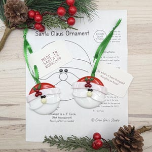 Pode incluir: Dois ornamentos de Natal redondos de vidro em forma de Pai Natal, com gorro vermelho, barba branca e detalhes de azevinho, pendurados por uma fita verde. Uma etiqueta diz Made in Santa's Workshop, a outra So, what's it been this year? Naughty or Nice!