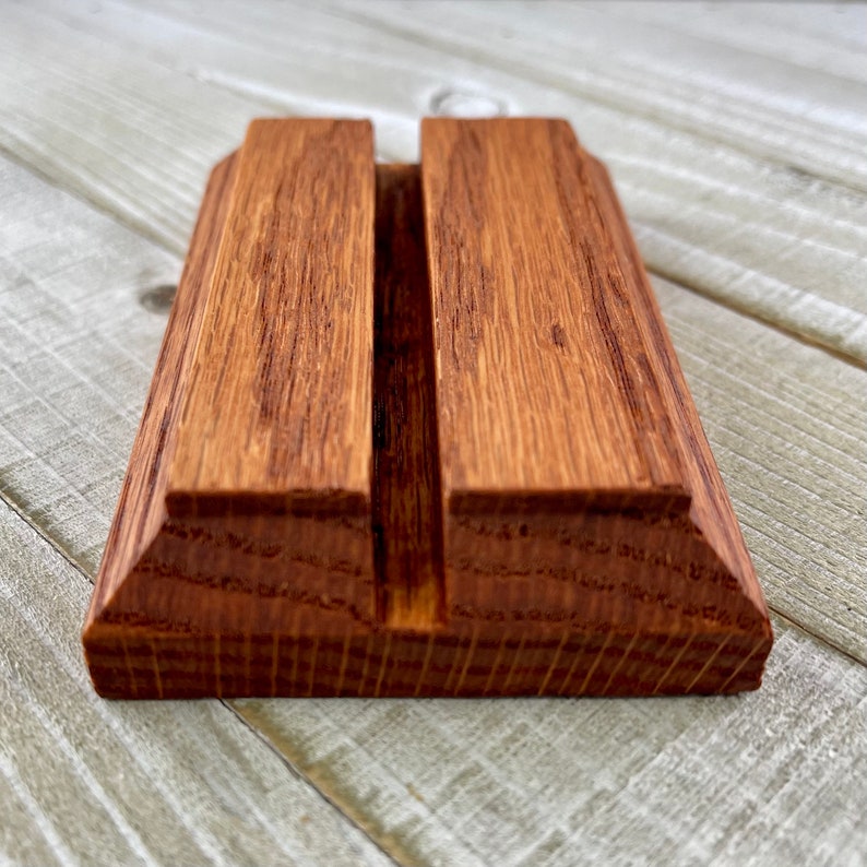 Op de afbeelding: Een houten telefoonstandaard met een warme, roodbruine kleur. De standaard heeft een rechthoekige basis met een verticale sleuf voor een telefoon of tablet. De houtnerf is zichtbaar, wat bijdraagt aan de natuurlijke uitstraling.