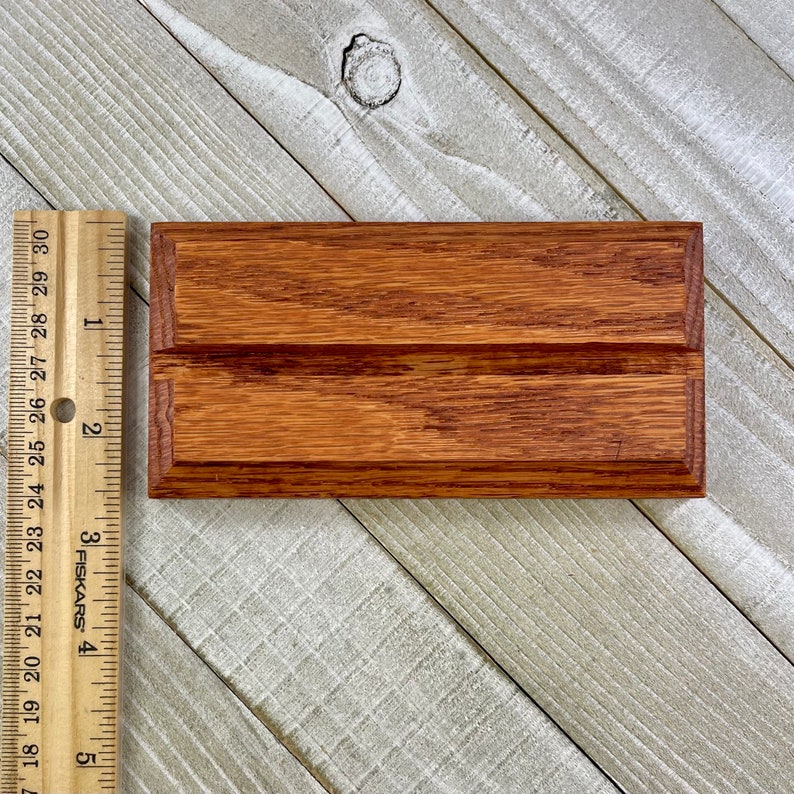 K&ouml;nnte beinhalten: Ein rechteckiger Visitenkartenhalter aus Holz mit einer satten, rotbraunen Oberfl&auml;che. Der Halter ist etwa 13 cm lang und verf&uuml;gt &uuml;ber einen gerillten Schlitz f&uuml;r Karten. Die Holzmaserung ist sichtbar und tr&auml;gt zu seiner nat&uuml;rlichen &Auml;sthetik bei.