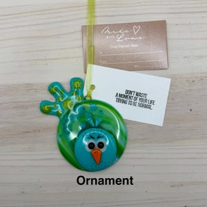 May include: A turquoise and green glass ornament in the shape of a bird with an orange beak and angry eyes. The ornament has a tag that says "Don't waste a moment of your life trying to be normal."