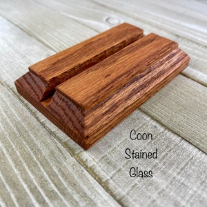 K&ouml;nnte beinhalten: Ein Holz-Handy-St&auml;nder mit einem satten, rotbraunen Farbton. Der St&auml;nder besteht aus zwei parallelen Holzst&uuml;cken mit einer Nut in der Mitte, um ein Telefon zu halten. Der Text "Coon Stained Glass" ist sichtbar.