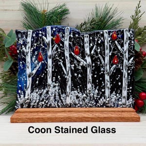 Op de afbeelding: Gebrandschilderd glaskunstwerk met een winterbos met witte berken, rode kardinalen en sneeuwval. Het glas is gemonteerd op een houten voet. De tekst "Coon Stained Glass" staat onderaan.