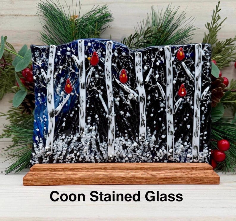 Op de afbeelding: Gebrandschilderd glaskunstwerk met een winterbos met witte berken, rode kardinalen en sneeuwval. Het glas is gemonteerd op een houten voet. De tekst "Coon Stained Glass" staat onderaan.