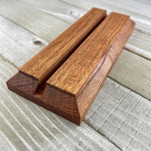 Op de afbeelding: Een houten telefoonstandaard met een natuurlijke houtnerf afwerking. De standaard is gemaakt van twee parallelle stukken hout die aan de basis zijn verbonden, met een sleuf om een telefoon of tablet vast te houden. Het hout heeft een warme, roodbruine tint.