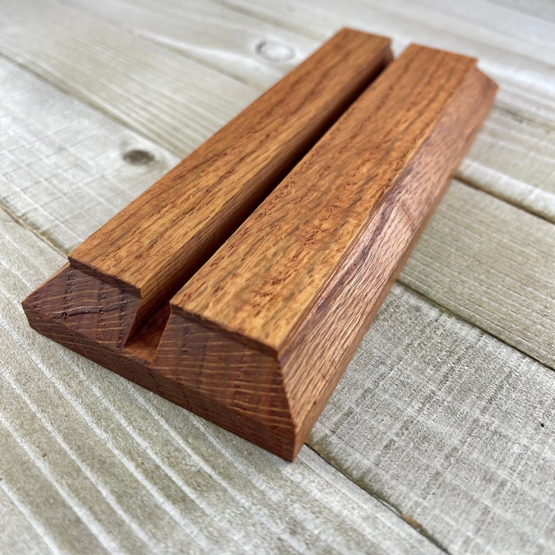Op de afbeelding: Een houten telefoonstandaard met een natuurlijke houtnerf afwerking. De standaard is gemaakt van twee parallelle stukken hout die aan de basis zijn verbonden, met een sleuf om een telefoon of tablet vast te houden. Het hout heeft een warme, roodbruine tint.