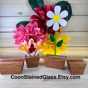 May include: Three stained glass pieces on wooden stands. The pieces are a white daisy with a yellow center, a red and white rooster, and a yellow flower. The stands are made of wood and have a natural finish.