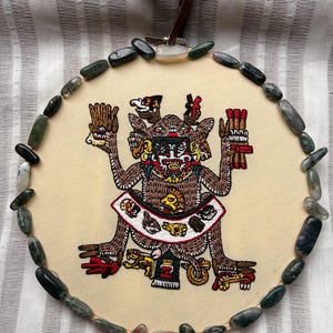 May include: Round, cream-colored embroidered wall hanging featuring a detailed illustration of a mythological figure with multiple arms and a decorative skirt. The artwork is framed with polished, multi-colored stones and a brown leather hanging strap.