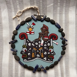 May include: Embroidered wall hanging featuring a stylized figure with red and black accents, set against a light blue background. The design is framed with dark stones and blue-grey crystals. A cream-colored cord is attached for hanging.