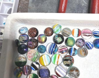 May include: A collection of small, round glass cabochons arranged in a white tray. The cabochons feature diverse patterns including iridescent swirls, metallic flecks, striped designs, and solid colours in shades of blue, green, brown, and pink.