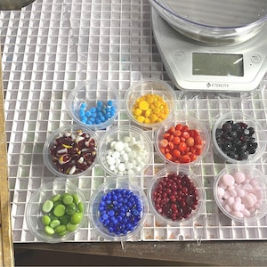 May include: Assortment of small glass beads in various colors, including red, white, blue, green, yellow, black, and pink, displayed in clear plastic containers on a white grid surface.