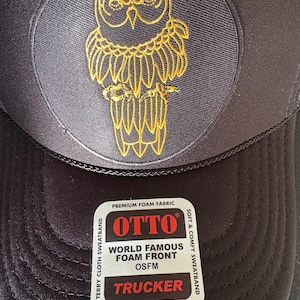 Op de afbeelding: Zwarte truckerpet met een cirkelvormige patch met een goud geborduurd uilontwerp. De pet heeft een schuimrubberen voorkant en een druksluiting. De patch leest "OTTO WORLD FAMOUS FOAM FRONT TRUCKER".