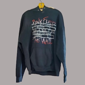 Puede incluir: Sudadera con capucha negra con bolsillo delantero, con el texto "Pink Floyd The Wall" en rojo y blanco sobre un diseño de pared de ladrillos. La sudadera está colgada de una percha amarilla.