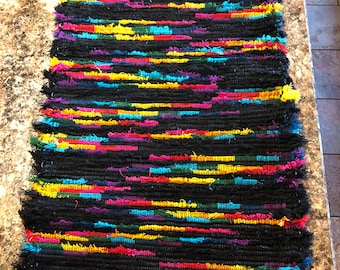 May include: A colorful rag rug with black, yellow, red, blue, green, and purple stripes. The rug has a fringe border.
