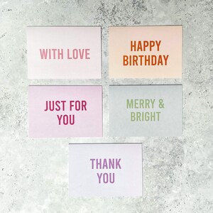 Puede incluir: Cinco tarjetas de felicitaci&oacute;n con diferentes mensajes. Las tarjetas son de color rosa, melocot&oacute;n, gris claro, morado claro y azul claro. Los mensajes son "With Love", "Happy Birthday", "Just For You", "Merry & Bright" y "Thank You".