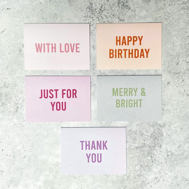 Puede incluir: Cinco tarjetas de felicitaci&oacute;n con diferentes mensajes. Las tarjetas son de color rosa, melocot&oacute;n, gris claro, morado claro y azul claro. Los mensajes son "With Love", "Happy Birthday", "Just For You", "Merry & Bright" y "Thank You".