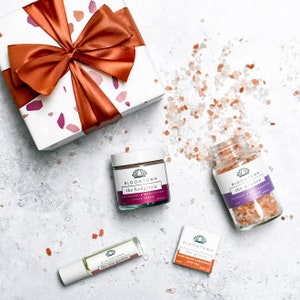 Puede incluir: Un set de regalo de productos para el ba&ntilde;o y el cuerpo de Bloomtown. El set incluye un exfoliante de az&uacute;car, sales de ba&ntilde;o y un aceite corporal. Los productos est&aacute;n envasados en frascos y botellas blancas con etiquetas moradas y rosas. Las etiquetas muestran el logotipo de Bloomtown y los nombres de los productos: "the hedgerow", "the meadow", "the rose garden" y "the grove".
