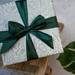 Puede incluir: Una caja de regalo blanca con un patr&oacute;n floral verde y un gran lazo de sat&eacute;n verde. La caja est&aacute; sobre una superficie de madera.