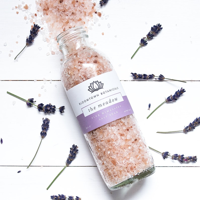 May include: A glass bottle filled with pink Himalayan salt soak, labeled "Bloomtown Botanicals the meadow Pink Himalayan Salt Soak". The bottle is surrounded by sprigs of lavender on a white wooden background.