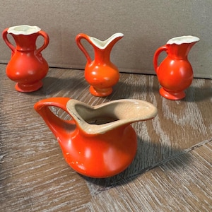 May include: A collection of four bright orange ceramic pitchers and vases. Three small vessels stand upright with handles and white rims, while one larger, wider pitcher with a prominent handle and a beige interior sits in the foreground.