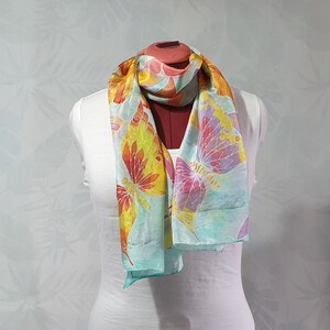 Può includere: Un foulard di seta blu chiaro con un motivo a farfalla in tonalità di arancio, giallo, rosa e viola. Il foulard è legato attorno a un manichino bianco.