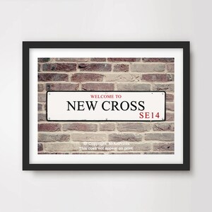 Op de afbeelding: Een wit straatnaambord met zwarte letters met de tekst "Welkom in New Cross SE14" tegen een achtergrond van een bakstenen muur.