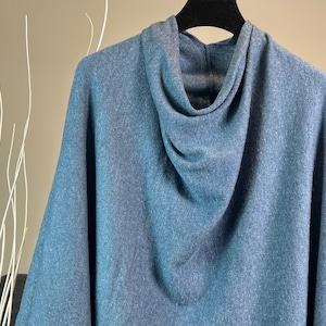 Puede incluir: Un poncho azul con capucha hecho de una tela suave de punto. El poncho tiene un cuello de pico y un ajuste holgado.