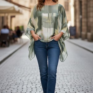 Puede incluir: Una mujer con una blusa estampada en verde y blanco y vaqueros azules. La blusa tiene un diseño suelto y fluido con cuello barco y mangas cortas. Los vaqueros son de color oscuro y la mujer lleva zapatos de esparto beige. El fondo es una calle adoquinada.