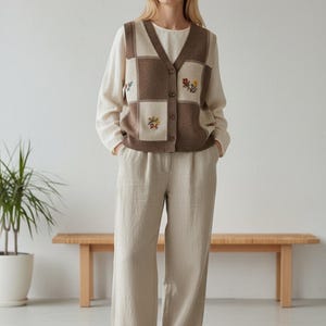 以下が含まれることがあります： ベージュの服装をした女性。ワイドパンツ、長袖シャツ、ボタン付きベストを着用しています。ベストは、茶色とクリーム色の正方形のパッチワークデザインで、花の刺繍が施されています。白いスニーカーでコーディネートされています。