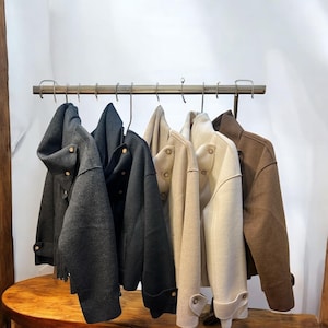 May include: A row of stylish jackets in various colors, including charcoal, black, beige, off-white, and brown, hang on a metal rack. Each jacket features a unique design with button details and a tailored fit, showcasing a modern aesthetic.