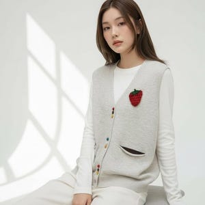 以下が含まれることがあります： 赤いイチゴのアップリケが付いた、ライトグレーのニットベスト。 ベストはVネック、カラフルなボタン、茶色のトリムが付いた小さなポケットが付いています。 白い長袖シャツの上に着用し、白いパンツと組み合わせています。