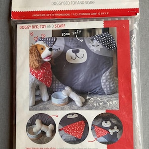 Può includere: Un modello di cucito per un letto per cani, un giocattolo a forma di osso e una bandana. Il modello include istruzioni per realizzare un letto per cani grigio e bianco con un naso e occhi neri, un giocattolo a forma di osso bianco e una bandana rossa con stampa di ossa bianche. Il modello è intitolato "Doggy Bed, Toy and Scarf" e include il testo "sooo soft" e "yum yum".
