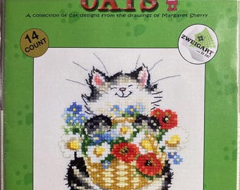 May include: Cross-stitch kit titled "CALENDAR CATS" featuring a smiling cat holding a basket of flowers. The kit includes a 14-count fabric and is designed by Margaret Sherry. The packaging is green with red text.
