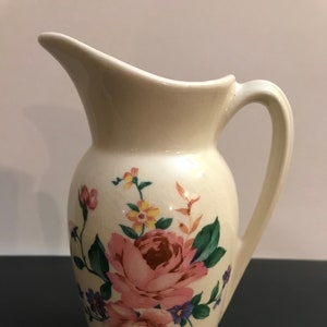 May include: A white ceramic pitcher with a floral design featuring pink, yellow, and purple flowers. The pitcher has a handle and a spout.