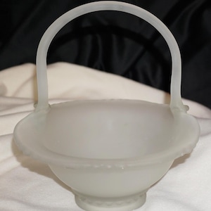 May include: A frosted glass bowl with a handle, shaped like a basket. The bowl is white and has a scalloped edge.