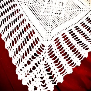 May include: Close-up of a white, handmade crochet doily. The intricate design features a geometric pattern with openwork details and a scalloped edge. The doily is placed on a dark red surface, highlighting the delicate texture.