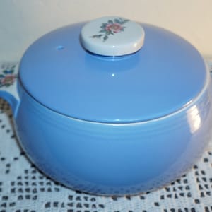 May include: A light blue ceramic casserole dish with a floral design on the lid and handles. The dish has a white rim and a white knob on the lid.