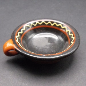 Puede incluir: Un pequeño cuenco de cerámica marrón con interior negro y borde amarillo y naranja. El cuenco tiene una sola asa.