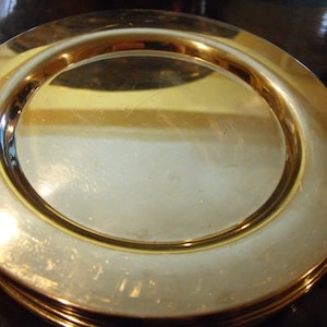 May include: A stack of gold-coloured plates with a shiny, reflective surface. Each plate has a wide, slightly raised rim and a smooth, flat centre. The plates appear to be made of metal and are stacked neatly on top of each other.