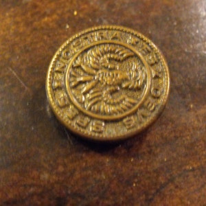 Może przedstawiać: Złoty metalowy guzik z wypukłym wzorem ptaka z rozpostartymi skrzydłami. Guzik ma napis "AUSTRIA" wokół krawędzi.