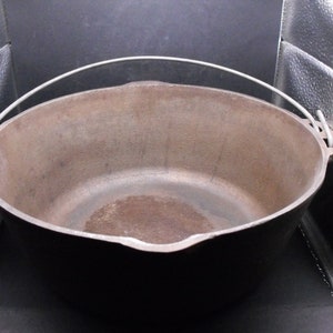 Puede incluir: Una olla grande de hierro fundido con un solo asa de plata. La olla está vacía y tiene un acabado rústico marrón.