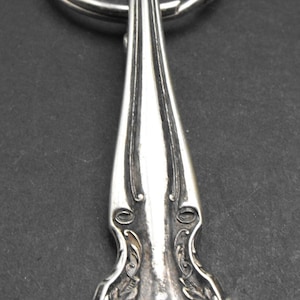 Puede incluir: Llavero de metal plateado con diseño de cuchara decorativa. La cuchara presenta detalles florales y de volutas ornamentados. El anillo del llavero está unido al mango de la cuchara. El fondo es gris oscuro.