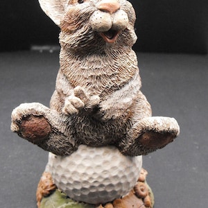 May include: A brown rabbit figurine sitting on a golf ball with the word "BOGEY" written on the base. The rabbit is holding a golf club in its paws.