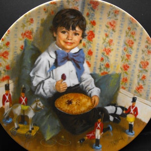 Könnte beinhalten: Ein Porzellanteller mit einer farbenfrohen Illustration eines jungen Jungen, der auf einem Kissen sitzt und eine Torte und einen Lutscher hält. Er ist umgeben von Spielzeugsoldaten und einem Spielzeugpferd mit Wagen. Der Teller hat einen floralen Musterrand.