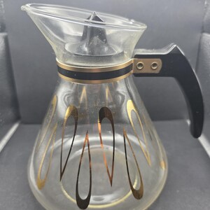 Op de afbeelding: Transparante glazen koffiekan met een zwarte handgreep en deksel. De kan heeft een gouden band en druppelvormige ontwerpen. De kan is leeg en lijkt vintage.