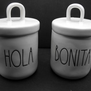 Puede incluir: Dos botes de cerámica blanca con tapas y asas. Los botes tienen letras negras. Un bote dice "Hola" y el otro dice "Bonita".