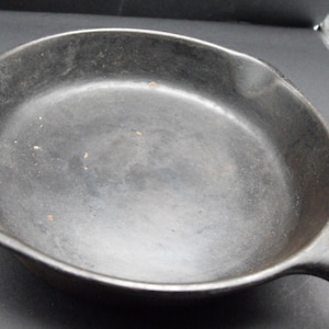 Könnte beinhalten: Eine schwarze Gusseisenpfanne mit einem Griff. Die Pfanne wird zum Kochen verwendet und hat eine abgenutzte, gut gewürzte Oberfläche.