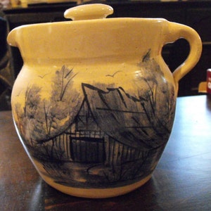 Könnte beinhalten: Eine cremefarbene Keramikkanne mit Deckel, die eine handgemalte Szene einer rustikalen Scheune in einem Waldgebiet zeigt. Die Scheune ist in Blautönen und Grautönen bemalt, mit einem dunkelgrauen Dach und einer Holztür. Die Bäume sind in Blautönen und Grautönen bemalt, mit einem hellblauen Himmel im Hintergrund.