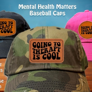 Puede incluir: Tres gorras de béisbol en negro, camuflaje y rosa, cada una con un parche de cuero que dice "GOING TO THERAPY IS COOL". La imagen también incluye el texto "Mental Health Matters Baseball Caps" y "8 Hat Colors To Choose From."