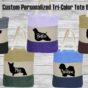 May include: Custom personalized tri-color tote bags with cream-colored handles. Each bag features a silhouette of a dog breed with a name. The bags are in various color combinations, including gray, black, purple, blue, and brown.